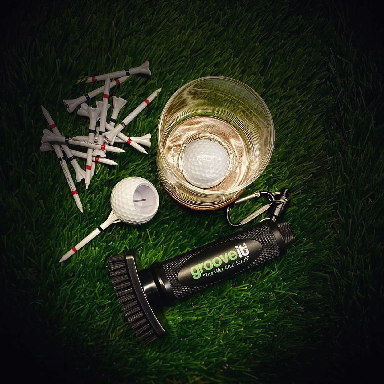 Grooveit Wet Club Scrub golf brush resting on turf with golf tees, a golf ball-shaped shot glass, and a drink with a floating golf ball ice cube.