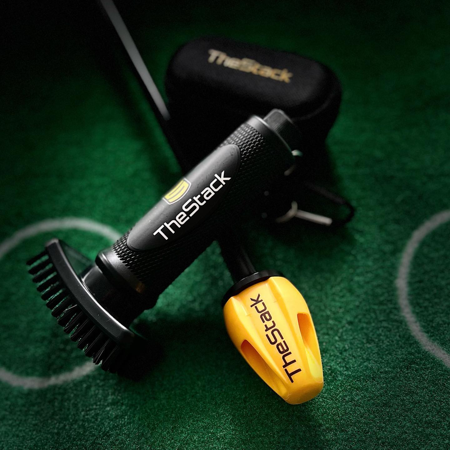 Close-up of a Grooveit brush custom-branded for TheStack, displayed on green turf next to a yellow TheStack training weight and black carrying case.