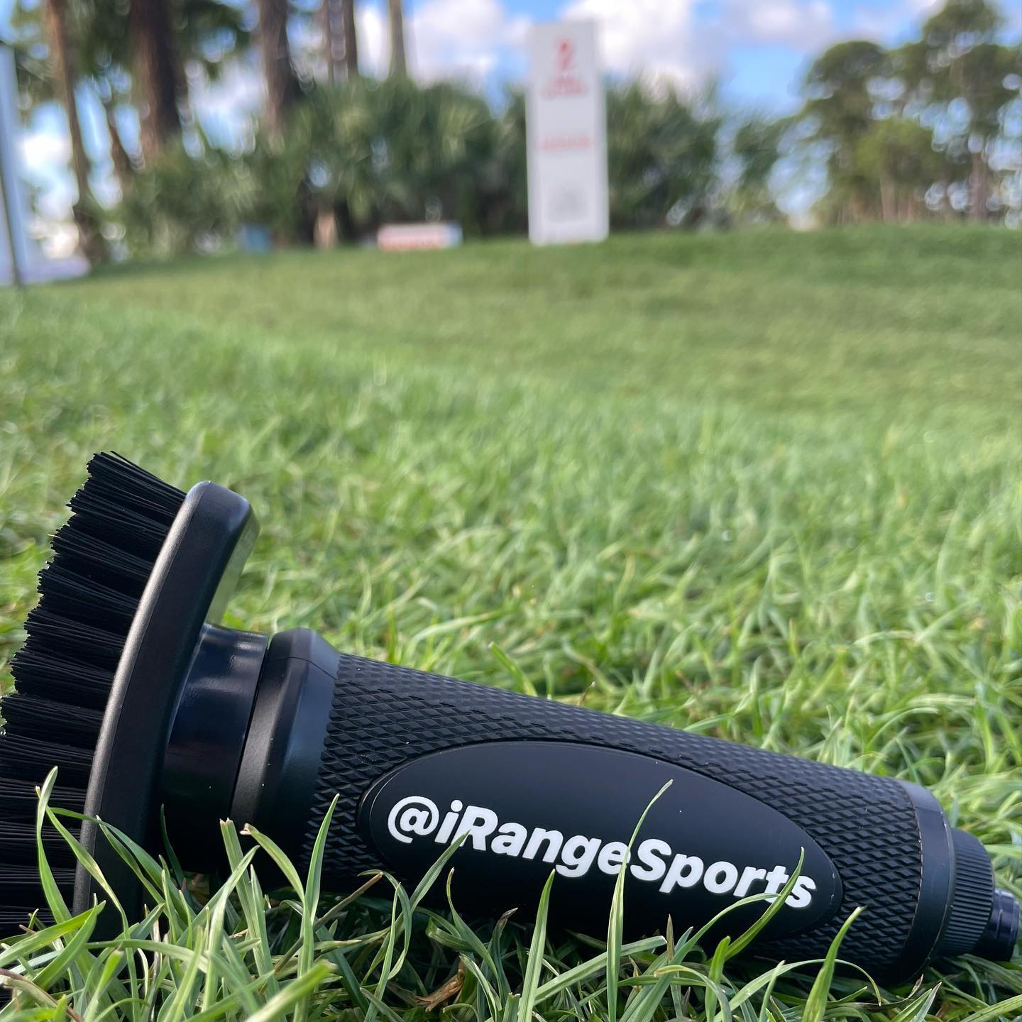 Grooveit golf brush customized with @iRangeSports logo lying on grass, with a golf course sign blurred in the background.