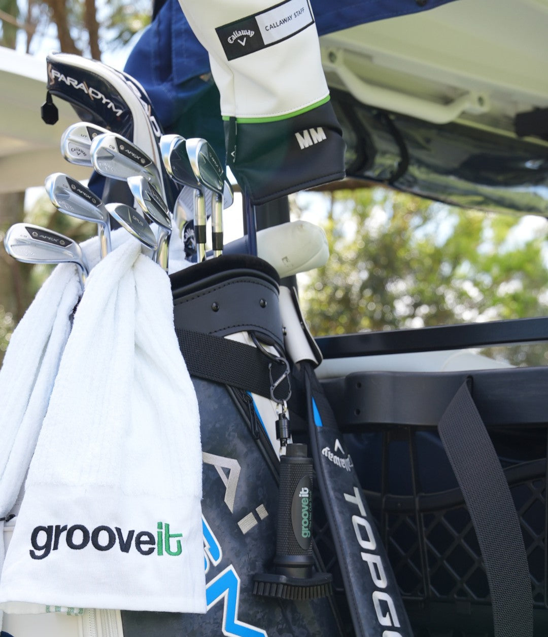 Close-up of a Callaway golf bag on a cart, featuring a Grooveit towel and Grooveit brush clipped to the side, surrounded by high-end golf clubs and accessories.
