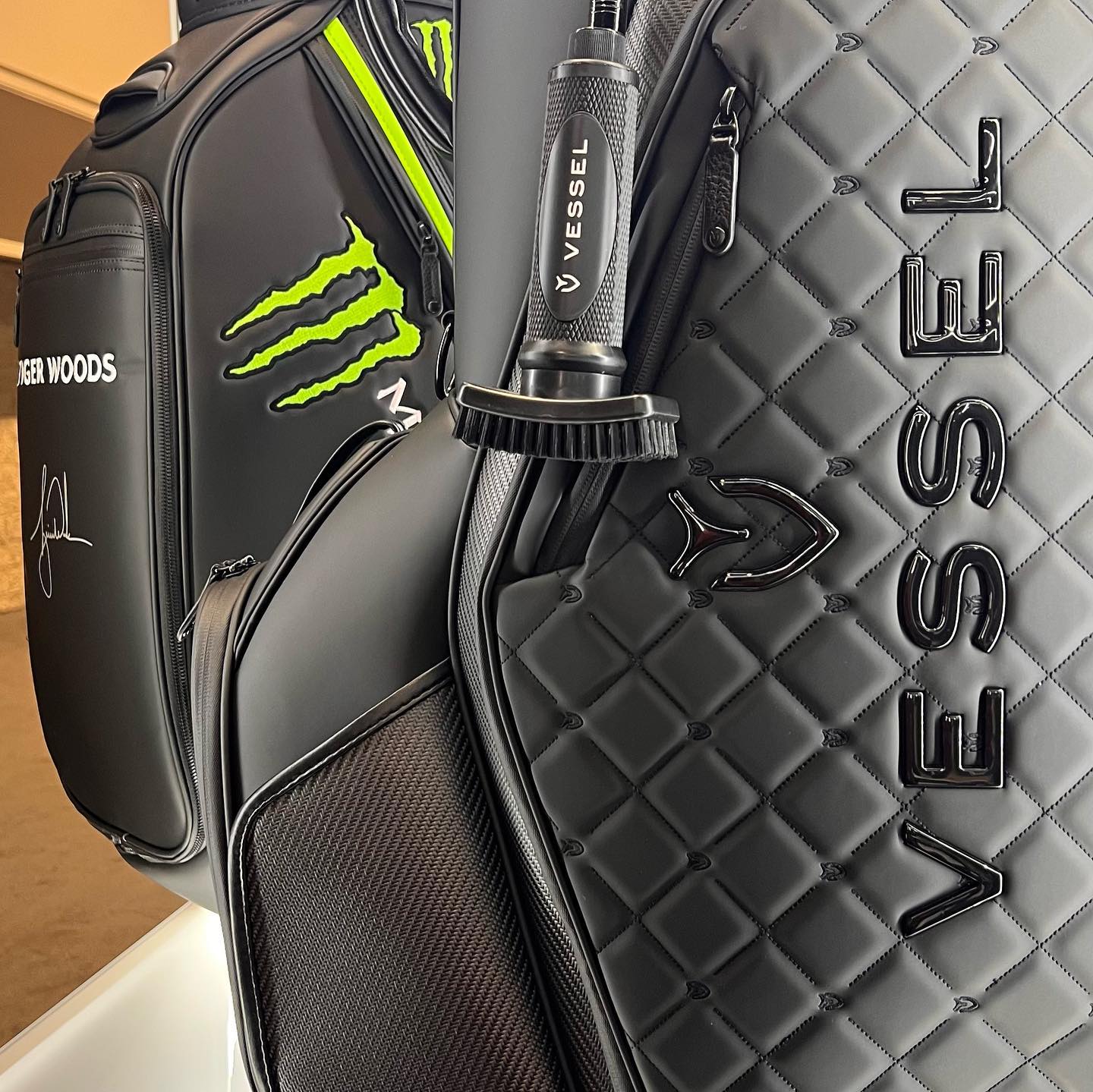Custom Grooveit golf brush with VESSEL logo clipped to a black quilted VESSEL golf bag, next to a Tiger Woods Monster Energy staff bag.