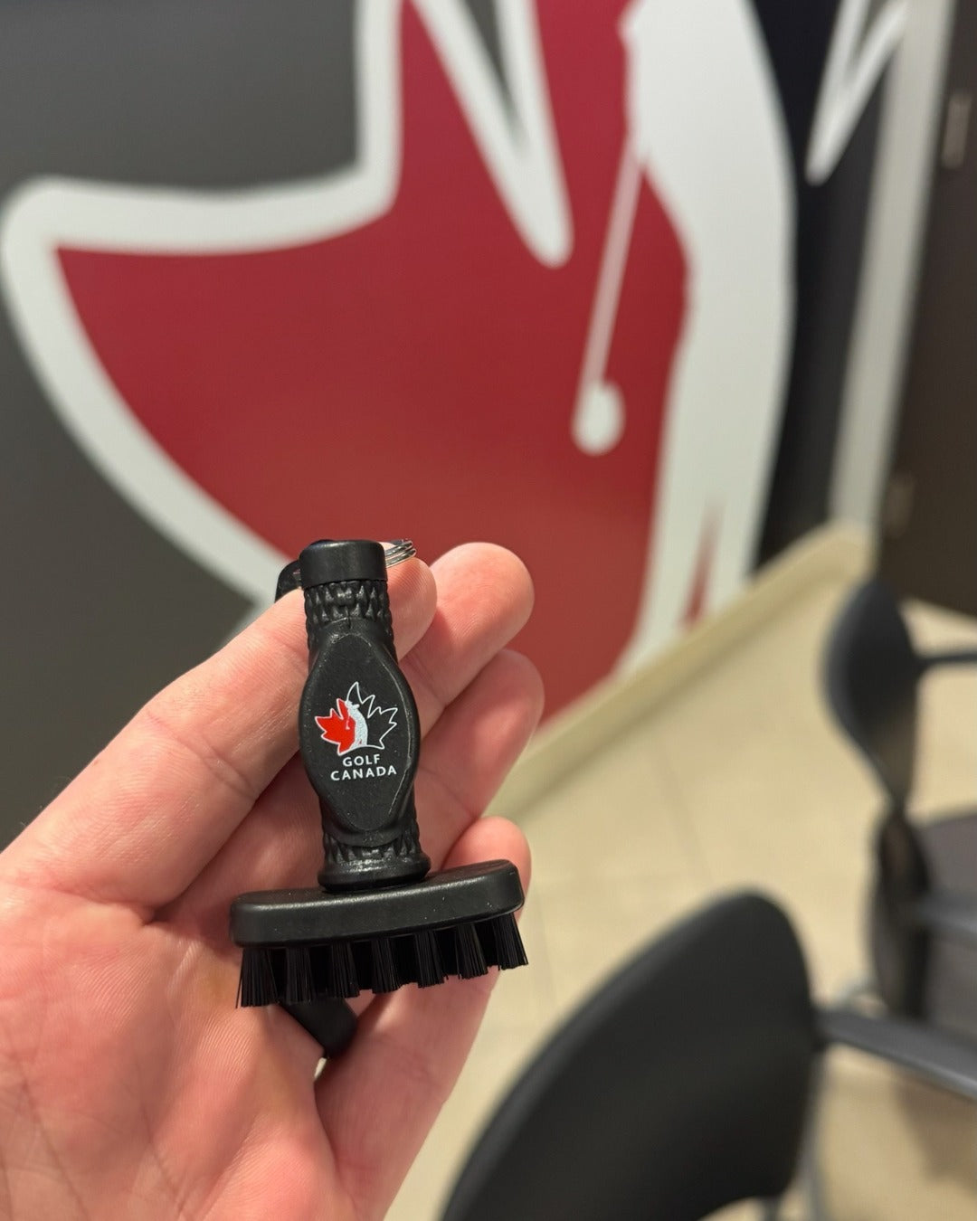 Hand holding a mini Grooveit brush customized with the Golf Canada logo, in front of a large red and white Golf Canada wall graphic.