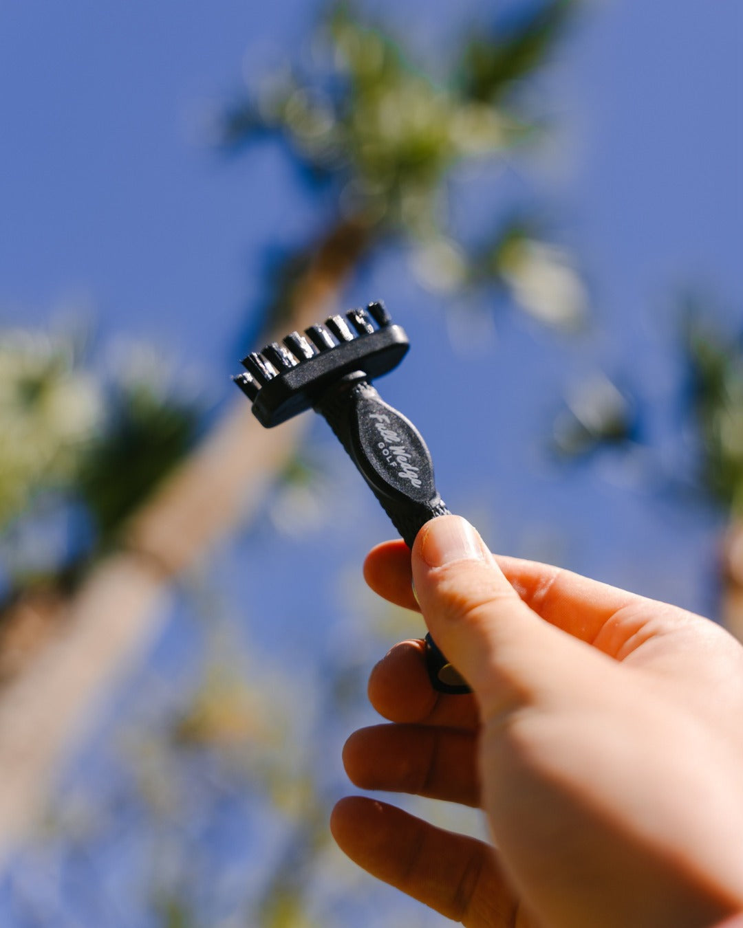 Hand holding a custom Grooveit miniG brush featuring the Fully Equipped Golf logo, with palm trees and a bright blue sky in the background.
