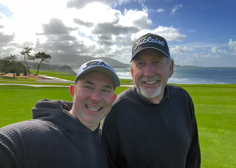 Two smiling men in Titleist hats posing for a selfie on a scenic golf course with the ocean, rolling hills, and dramatic clouds in the background.