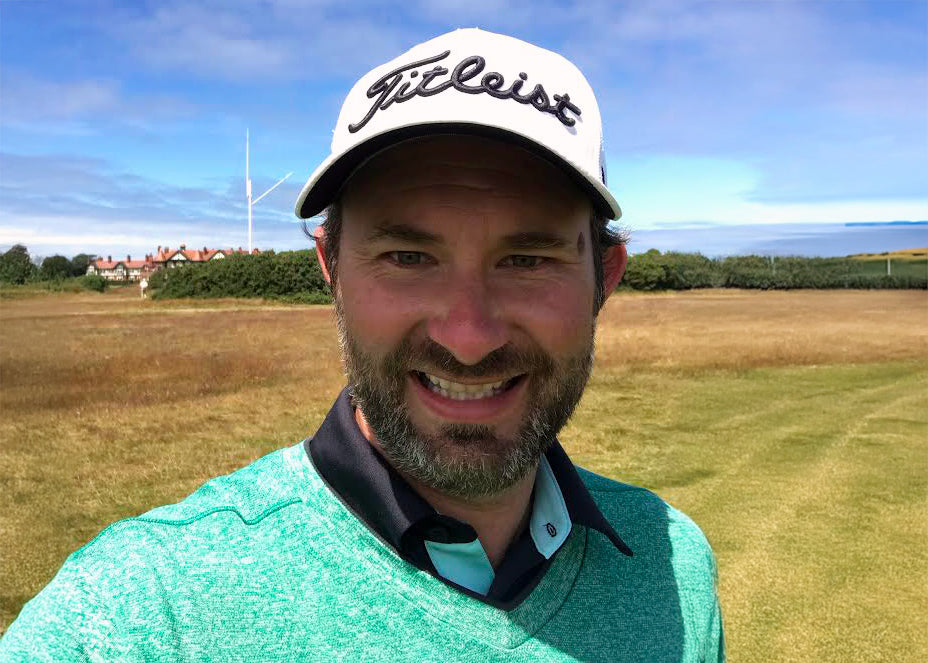 Andrew smiling on a sunny golf course wearing a green shirt and a white Titleist hat, with ocean and coastal buildings in the background.