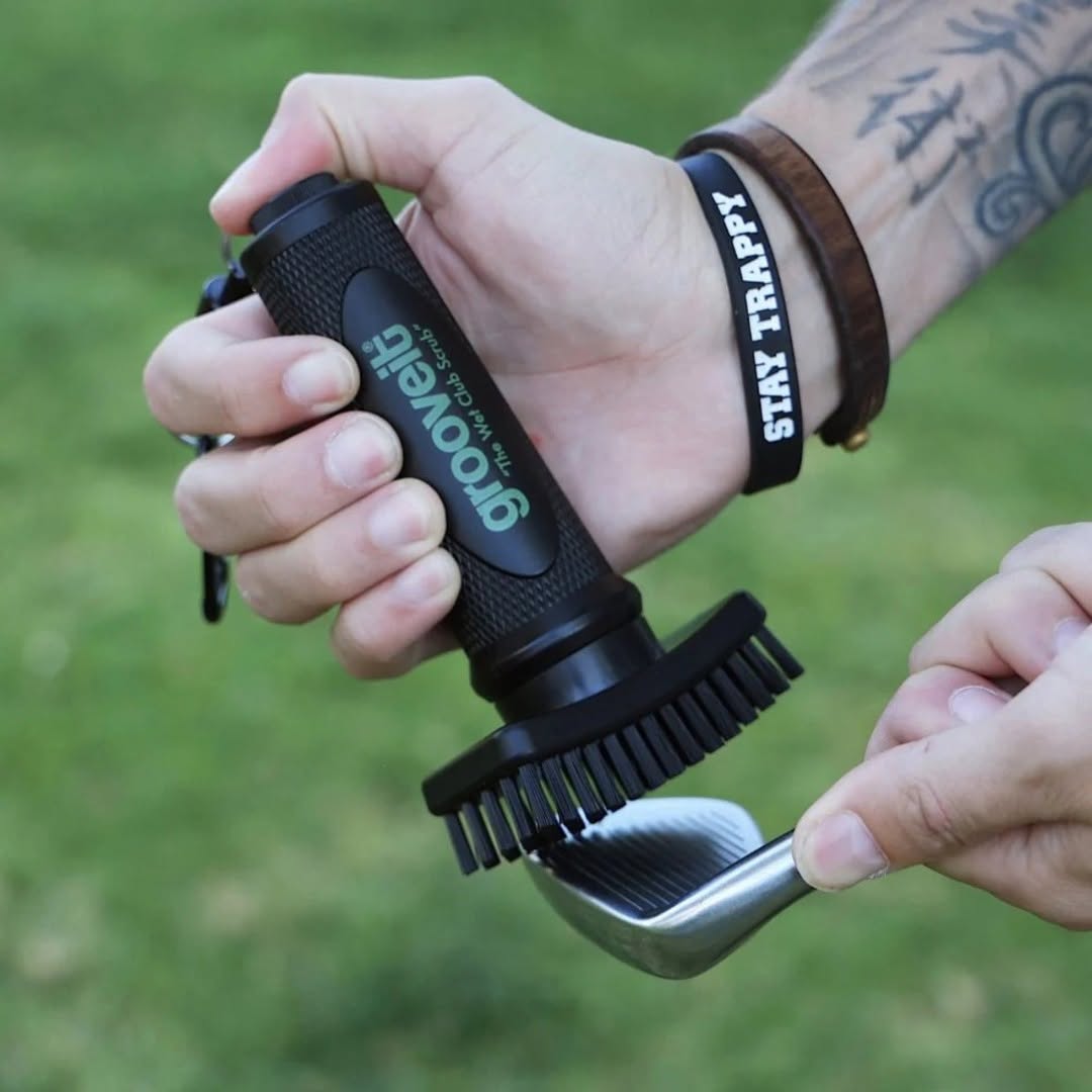 Close-up of a person using a Grooveit “Wet Club Scrub” golf brush to clean the grooves of an iron club, with visible wristbands and tattoos.