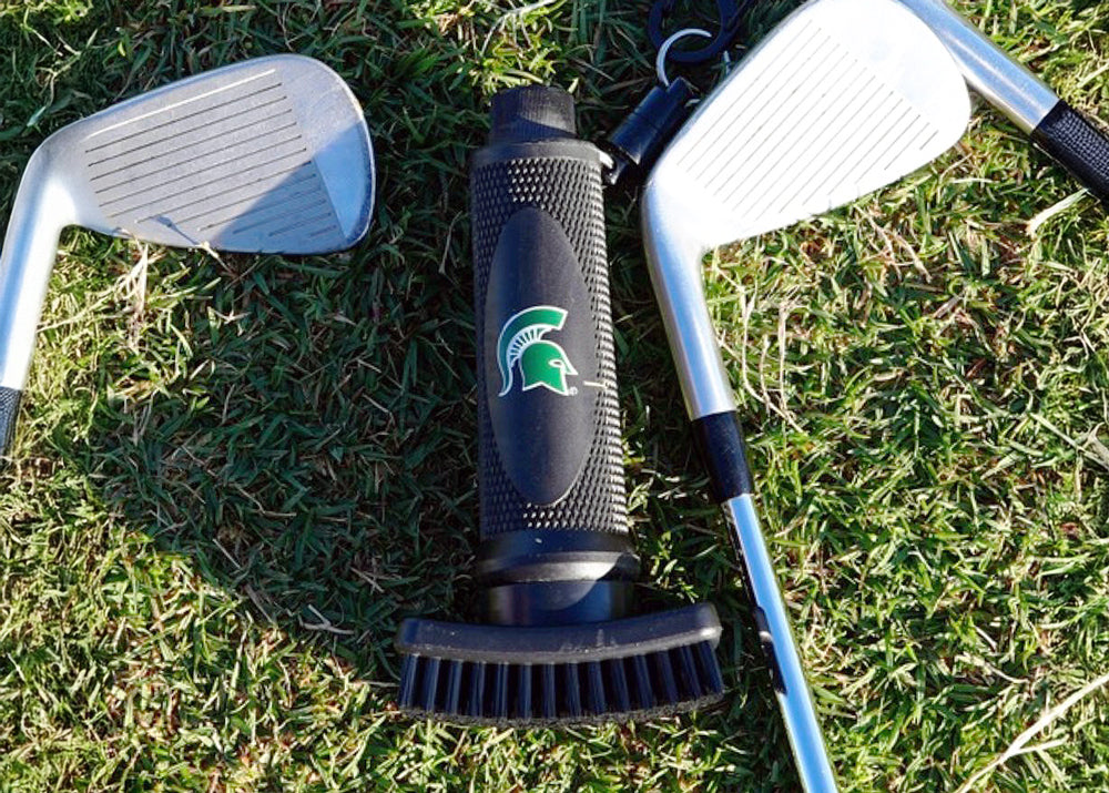 Grooveit golf club cleaning brush with the Michigan State Spartans logo, placed on grass between two golf irons.