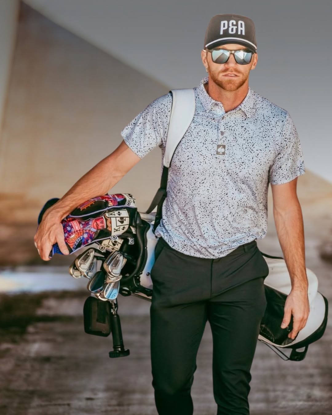 Man wearing sunglasses and a P&A hat walking with a golf bag over his shoulder, with a Grooveit brush visible hanging from the clubs.