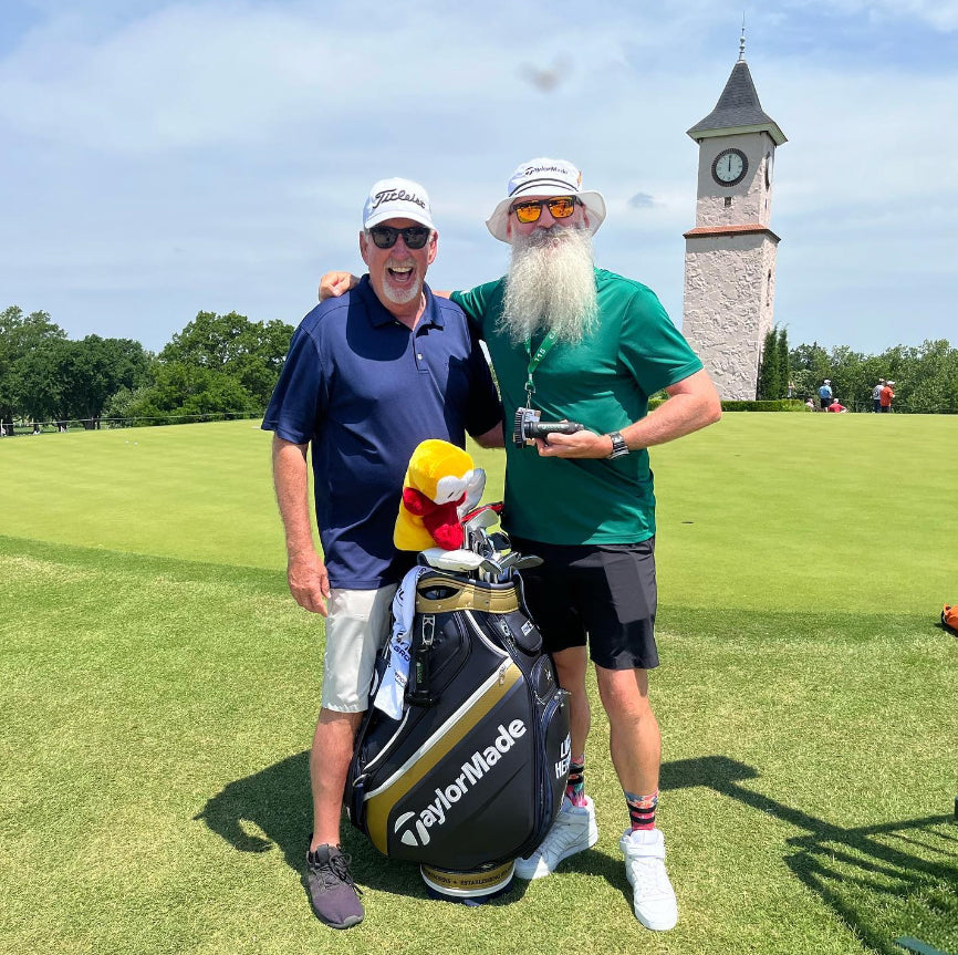 Two men smiling on a golf course next to a TaylorMade golf bag, with one holding a Grooveit Brush. A clock tower and other golfers are visible in the background.
