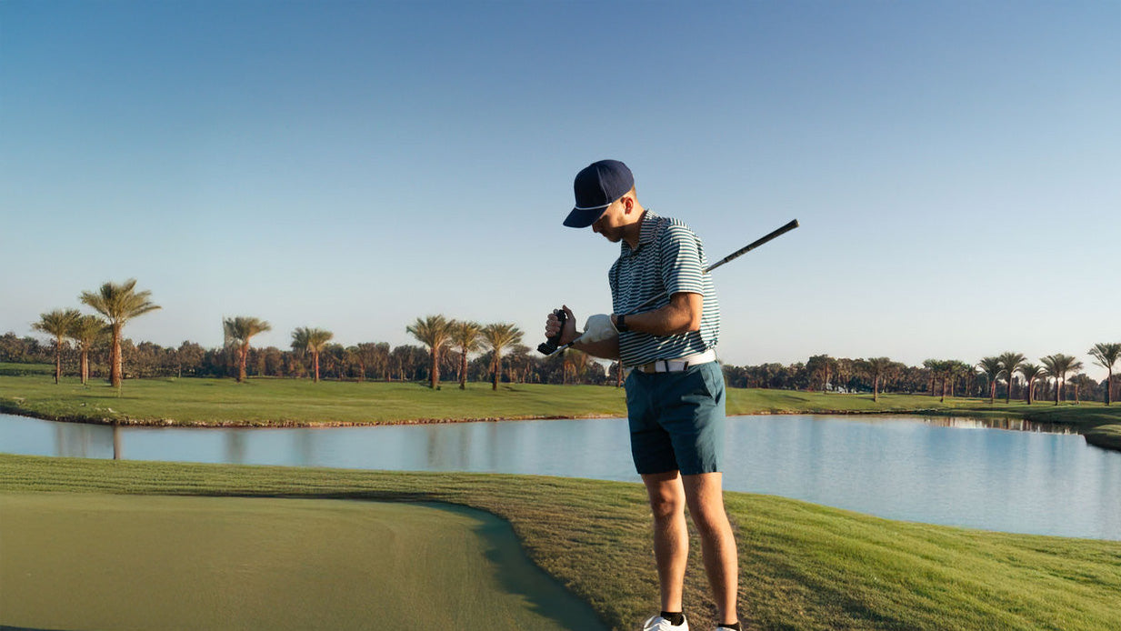 Golfer standing near a green by a lake, cleaning a golf club with the Grooveit brush, on a palm-lined course during a sunny day.