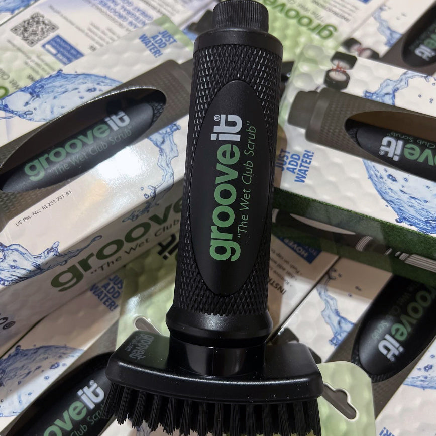 Close-up of a Grooveit "Wet Club Scrub" golf brush surrounded by retail packaging and boxes, highlighting product branding and bristle design.