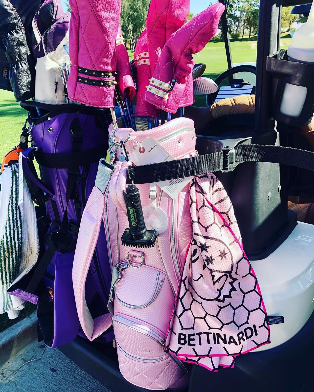 Pink golf bag with studded pink headcovers, a Grooveit brush, and a Bettinardi towel attached, sitting on a golf cart next to a purple bag.