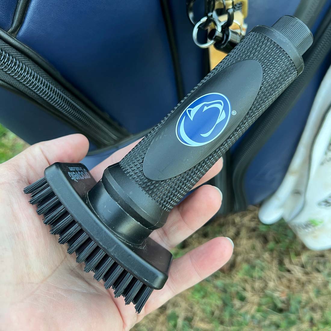Hand holding a Grooveit golf club cleaning brush with the Penn State Nittany Lions logo on the handle, attached to a golf bag in the background.
