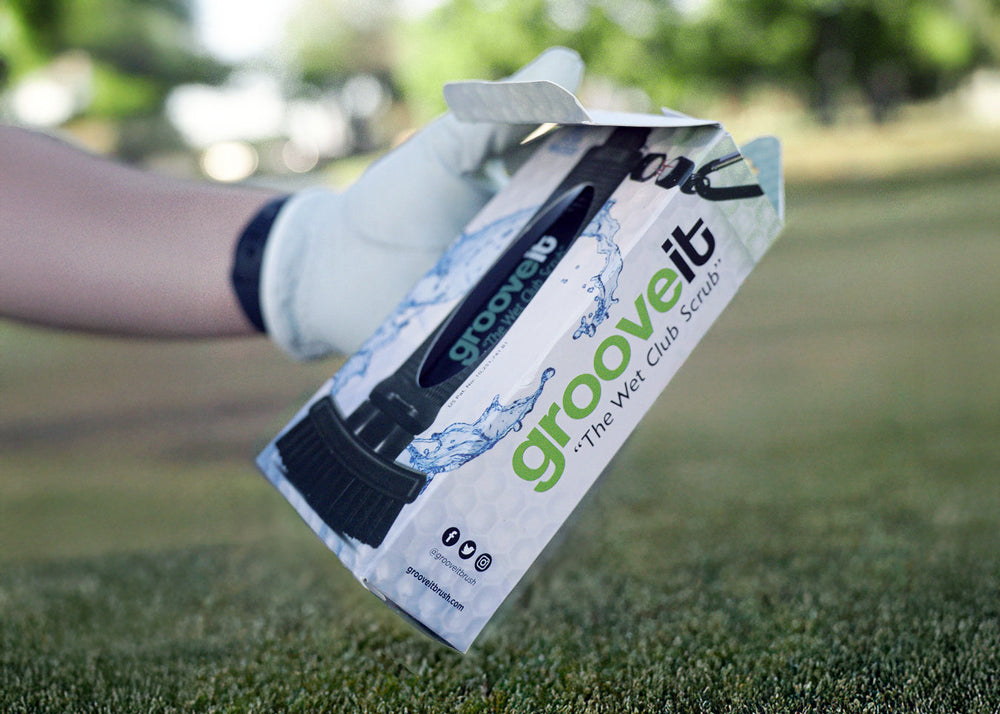 A gloved hand holding a packaged Grooveit “Wet Club Scrub” brush on a golf course.