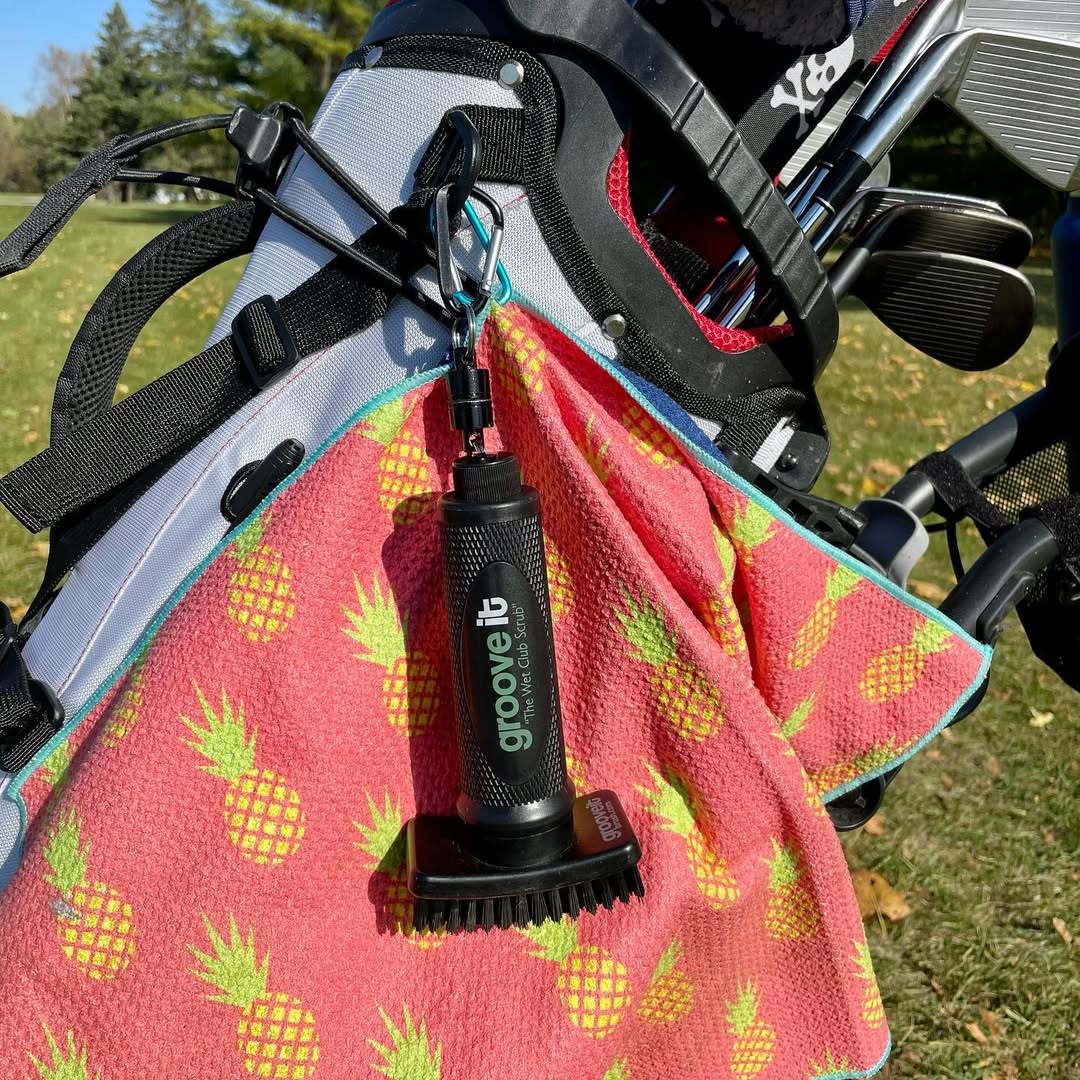 Grooveit golf brush clipped to a golf bag, hanging over a bright pink towel with yellow pineapples, with clubs and green grass in the background.