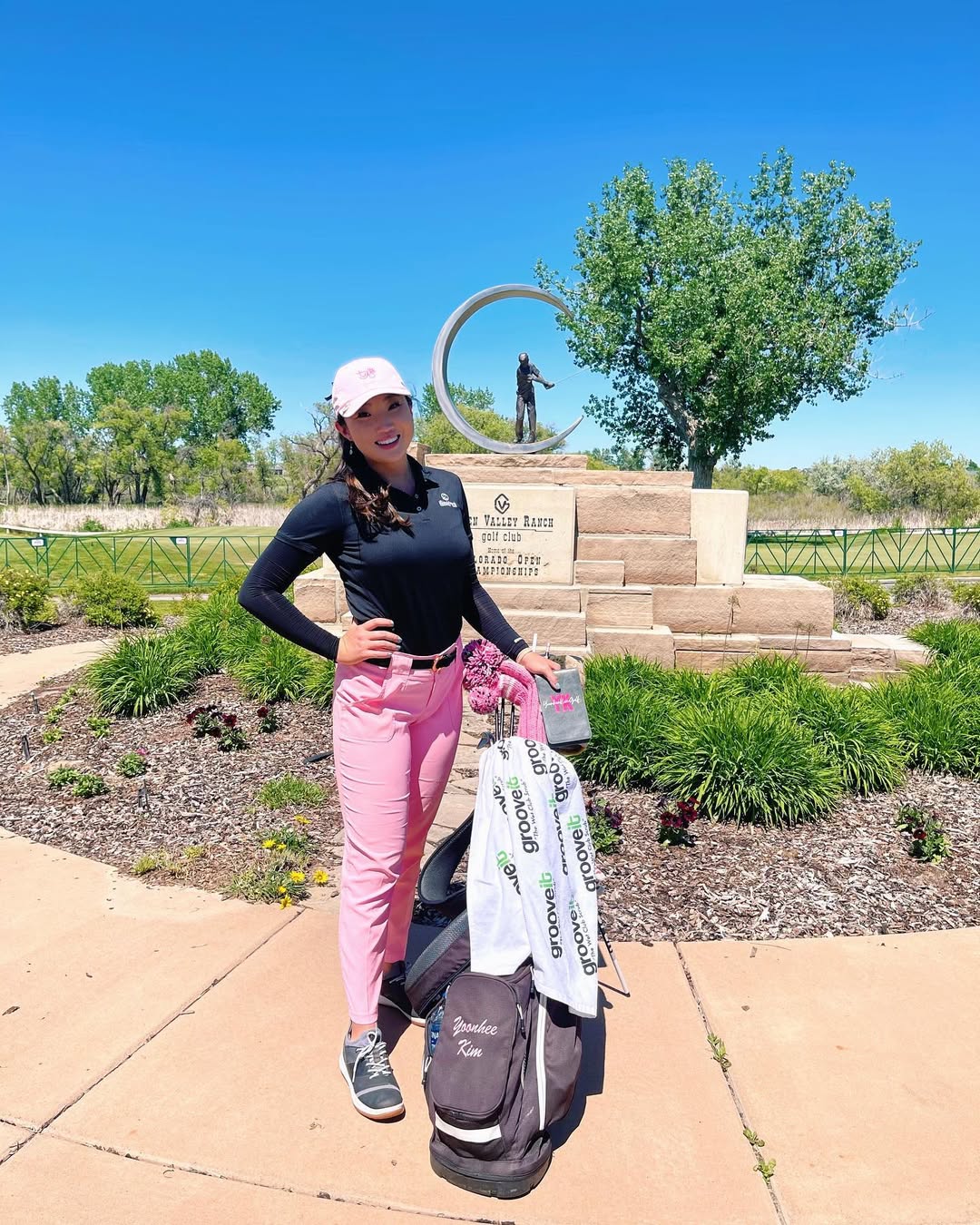 Female golfer smiling next to her golf bag with a Grooveit towel draped over it, standing in front of a stone statue at a sunny golf course.