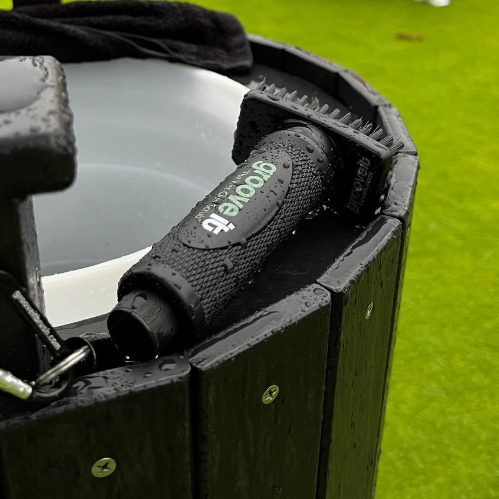 Grooveit “The Wet Club Scrub” golf brush resting on a wet ball washer station, covered in rain droplets on a green practice area.