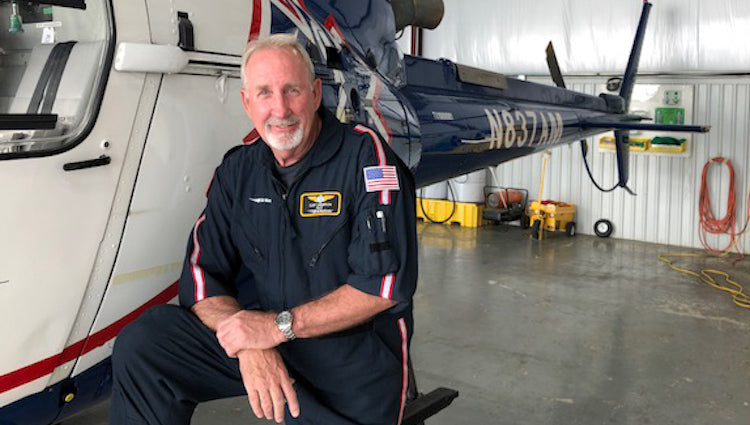 Clint Sanderson in a flight suit with American flag patch sitting next to a blue and white medical helicopter inside a hangar, smiling at the camera.