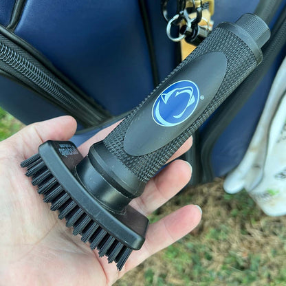 Hand holding a Grooveit golf club cleaning brush with the Penn State Nittany Lions logo on the handle, attached to a golf bag in the background.